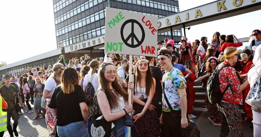 Grupa studentów ubranych w stylu kultury hippisowskiej stoi przed rektoratem. Dziewczyna na środku kadru trzyma transparent z napisem 