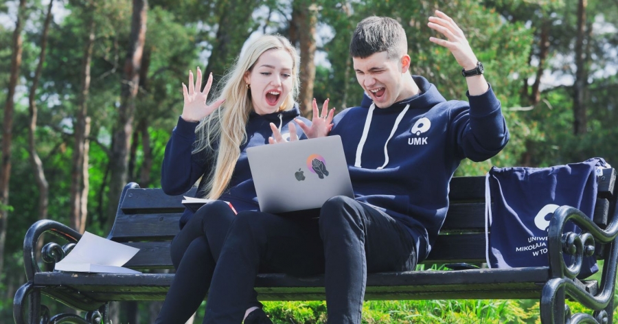 Dwoje studentów siedzi na ławce, mają na sobie granatowe bluzy z logo UMK. Student trzyma laptop na kolanach. Dziewczyna i chłopak mają uniesione ręce i otwarte usta. W tle rosną drzewa z zielonymi liśćmi