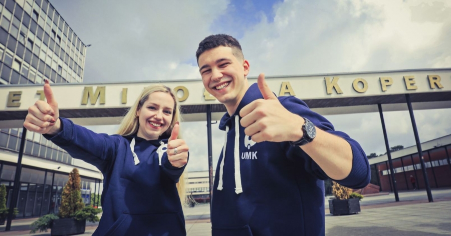 Dziewczyna i chłopak w bluzach z logo UMK pokazują uniesione kciuki przed budynkiem rektoratu