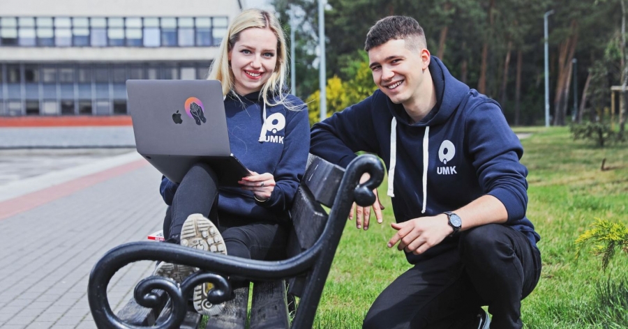 Dwoje studentów pozujących do zdjęcia w miasteczku uniwersyteckim. Dziewczyna trzyma na kolanach otwarty laptop