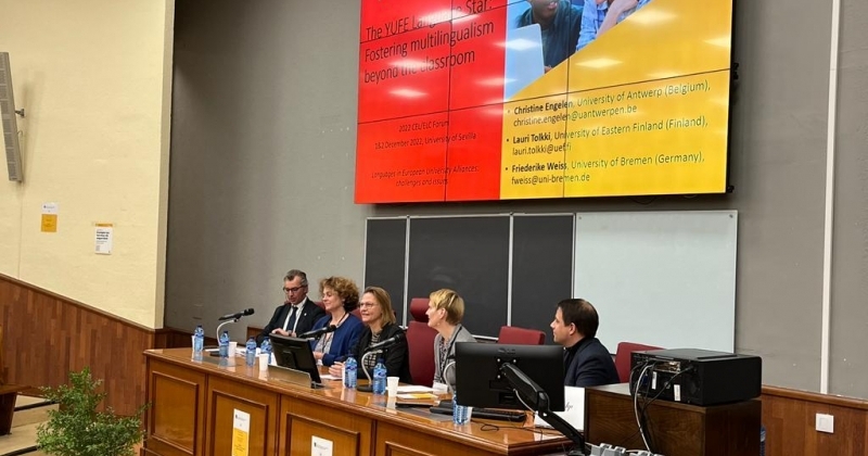 Five people are sitting at a lecture desk, a screen above them shows one presentation slide