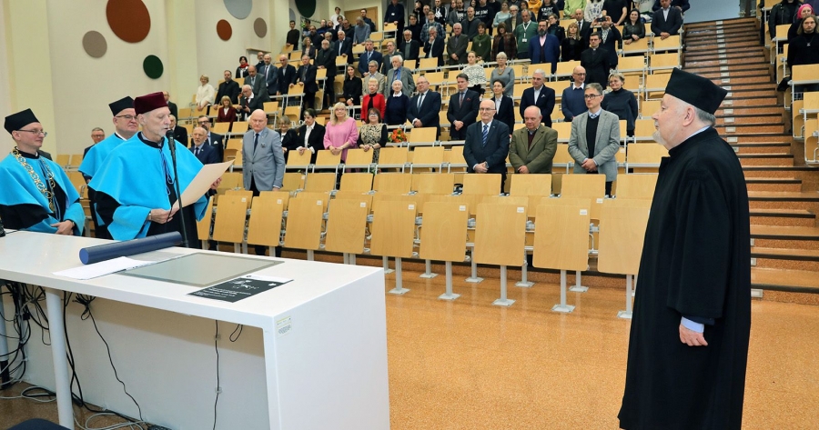 Starszy mężczyzna z brodą w czarnej todze i birecie stoi w sali audytoryjnej naprzeciw trzech mężczyzn, z których jeden odczytuje tekst dyplomu; w tle stojąca publiczność