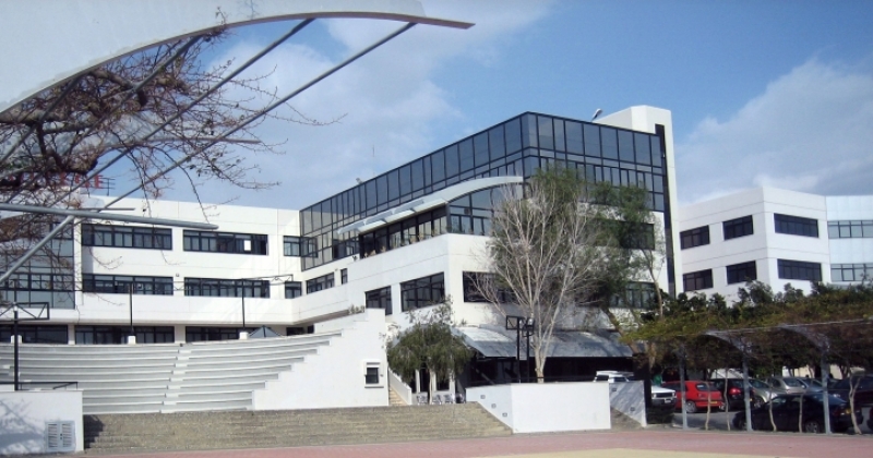 In the foreground, the amphitheater is visible against the background of the university building. The amphitheater and the building are made in a modern style, the white walls contrast with the glazed fragments of the façade. Trees and bushes, a fragment of the parking lot are also visible on the right side. Photo taken in full sun on a cloudless day, blue sky visible at the top of the frame. 