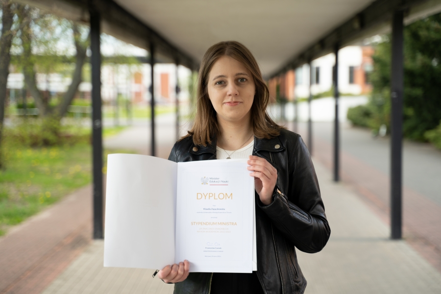 Zdjęcie portretowe kobiety, trzymającej w dłoniach otwartą teczkę z informacją o przyznanym stypendium Ministra. Kobieta stoi na rozmytym tle budynku Wydziału Nauk Ekonomicznych i Zarządzania UMK w Toruniu