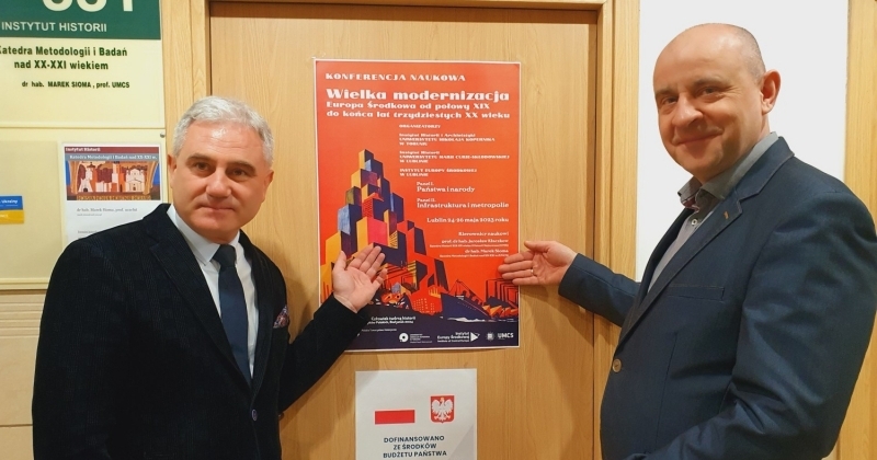 Dwóch mężczyzn rękoma wskazuje wiszący na drzwiach plakat informujący o konferencji.