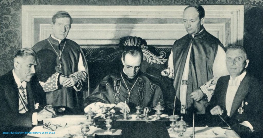 Papież przy stole podpisuje dokumenty, wokół stoi dwóch kapłanów i siedzą dwaj przedstawiciele Niemiec