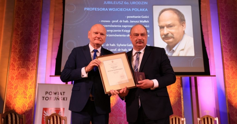 Prezydent Zaleski i prof. Polak pozują do zdjęcia po wręczeniu medalu. Prof. Polak w lewej dłoni trzyma medal, a prawą podtrzymuje dyplom w ramce. W tle widać wyświetlony program wydarzenia