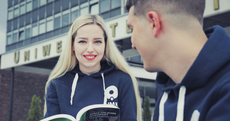 Dziewczyna trzyma książkę i patrzy w obiektyw. Obok niej siedzi chłopak i patrzy na książkę. Za nimi widać fragment rektoratu i napisu Uniwersytet Mikołaja Kopernika