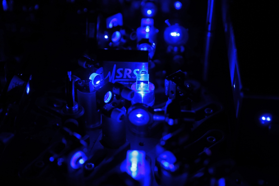 Fragment układu laserowego optycznego zegara atomowego. Czarne tło. Elementy układu świecą ciemnym niebeiskim światłem 
