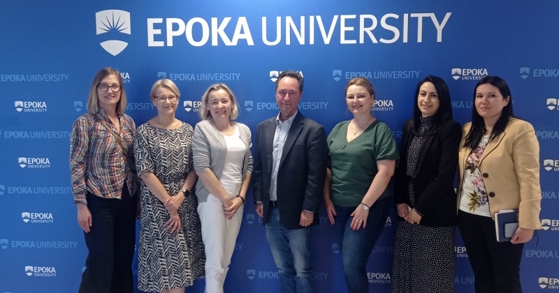 Na tle dużego baneru z nazwą i logo Uniwersytetu Uniwersytetu Epoka w Tiranie pozują uczestnicy spotkania, którzy są nauczycielami akademickimi obu uniwersytetów. Od lewej: mgr Joanna Ososińska-Ciechomska, mgr Ewa Kowalska, mgr Hanna Łysiak, prof. dr David Felsen, mgr Katarzyna Gałka, M.A. Ada Cara, Ph.D. Alba Gerdeci.