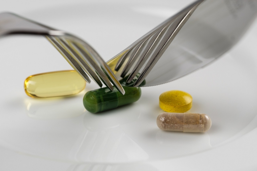 Różnokolorowe tabletki w podłużnym kształcie, jedna okrągła. Do zielonej, podłużnej, tabletki są przyłożone: widelec i nóż w takim ułożeniu jak podczas jedzenia posiłku.