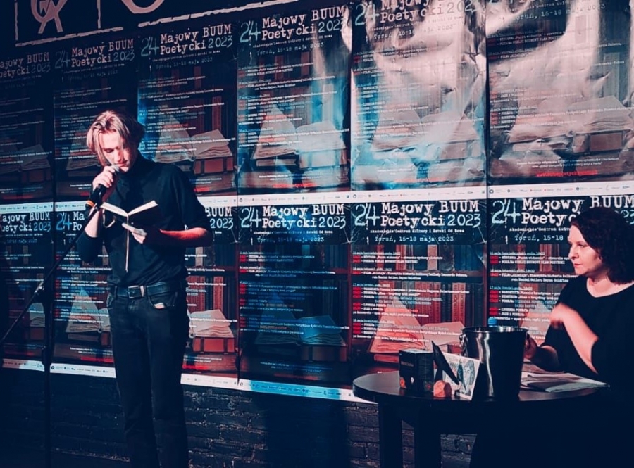 Fragment sceny. Z prawej strony młoda kobieta siedząca przy stoliku (ubrana w ciemną bluzkę). Z lewej strony, na scenie, stoi młody mężczyzna (ubrany na czarno), który czyta książkę. W tle plakaty pt. 24 Majowy BUUM Poetycki 2023.