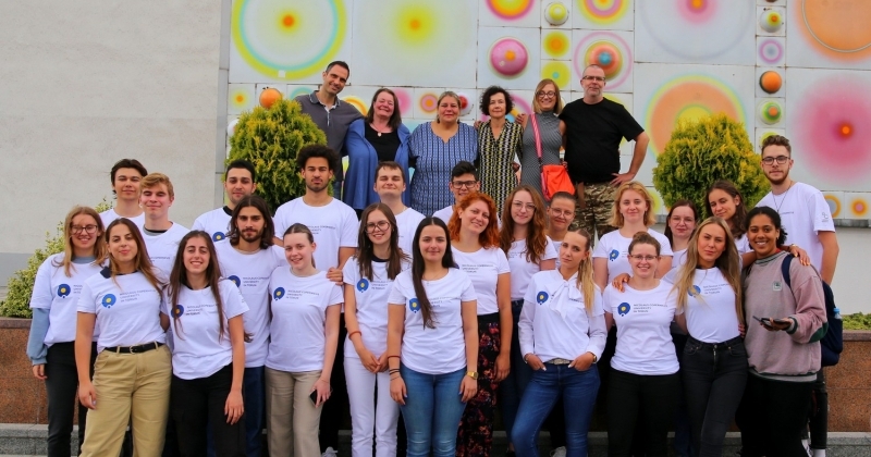na pierwszym planie uśmiechnięci studenci w dwóch rzędach ubrani w białe t-shirty z logo UMK. W trzecim rzędzie wykładowcy. Zdjęcie zrobione na tle dekoracyjnej fasady Auli UMK z systemem planet. 