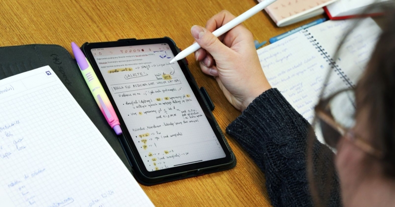 Zdjęcie zrobione znad głowy osoby uczącej się języka obcego. Na stole widać tablet z notatkami, obok otwarte zeszyty i zapisane kartki. Widać również rękę trzymającą długopis do tableta