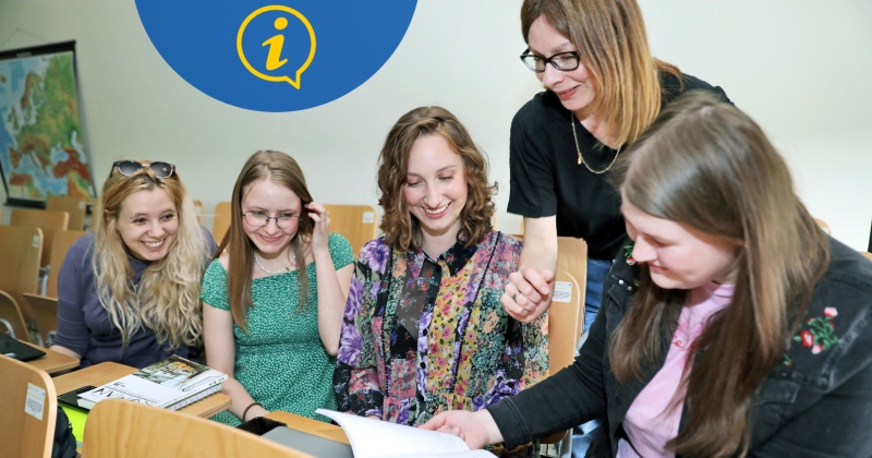 W sali wykładowej nauczycielka pochyla się nad czterema studentkami. Każda ze studentek ma przed sobą zeszyty ćwiczeń. 
