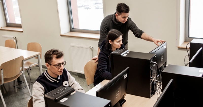 Dwójka studentów siedzi przed monitorami, wykładowca stoi i coś im pokazuje