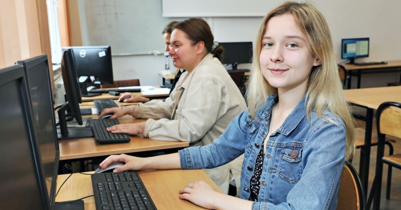 Kilka osób siedzi w sali wykładowej przy komputerach, dziewczyna najbliższej patrzy w obiektyw i się uśmiecha