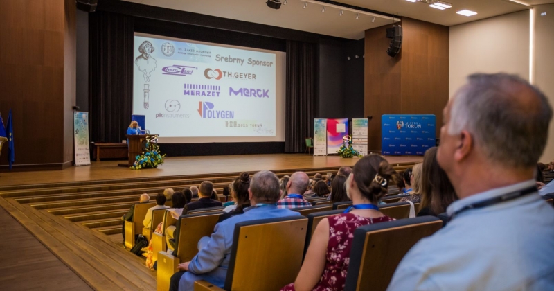 Aula UMK, ludzie na widowni, na scenie przy pulpicie przemawia jedna osoba, w tle wyświetlana jest prezentacja