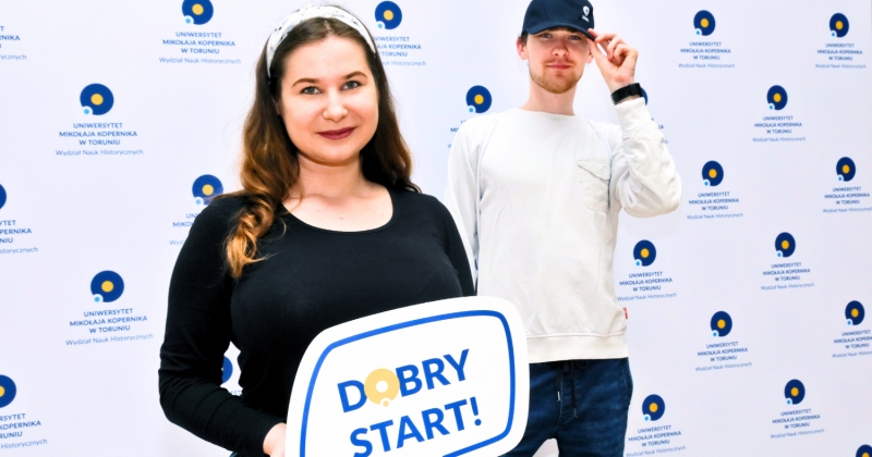 Dziewczyna i chłopak w czapce z daszkiem stoją przed ścianką z logo WNH. Dziewczyna ma napis "Dobry Start!"