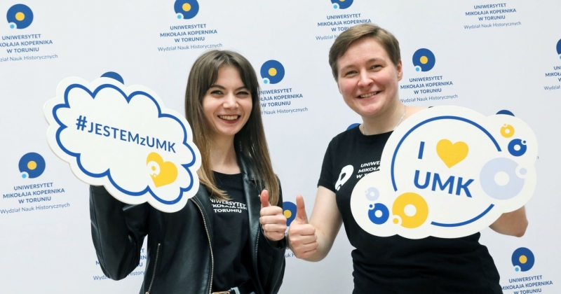 Dwie osoby na tle ścianki z logo UMK trzymają tabliczki "jestem z UMK" oraz "I love UMK" i pokazują kciuki podniesione w górę
