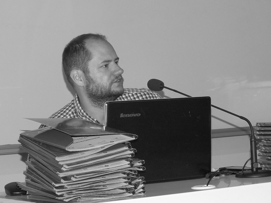 Młody mężczyzna z krótką brodą, widoczny z profilu, za laptopem i stosem teczek, wygłasza wykład w auli Collegium Humanisticum UMK.