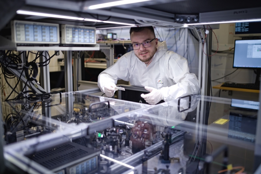 Na zdjęciu widzimy osobę pracującą w laboratorium. Osoba ta ma na sobie biały fartuch laboratoryjny i rękawice. Pracuje przy dużym urządzeniu z wieloma przewodami i kablami, które wygląda na komputer lub serwer. W ręku trzyma małe czarne urządzenie. W tle widoczne są różne sprzęty laboratoryjne.