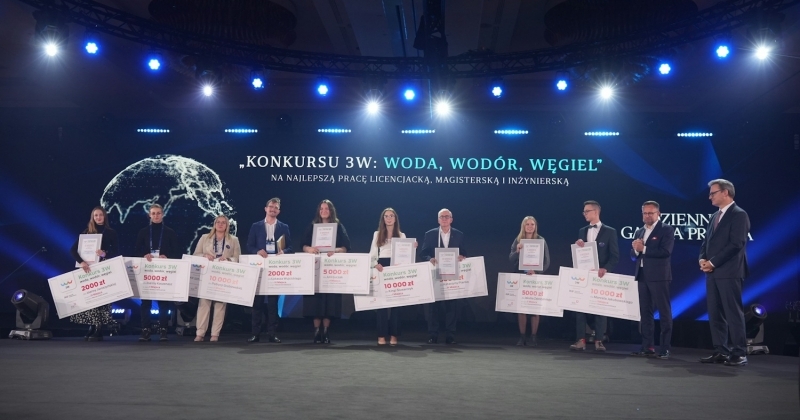 Dziewięcioro laureatów konkursu 3w stojący z duzymi czekami obok wręczającego nagrody w konkursie prac dyplomowych