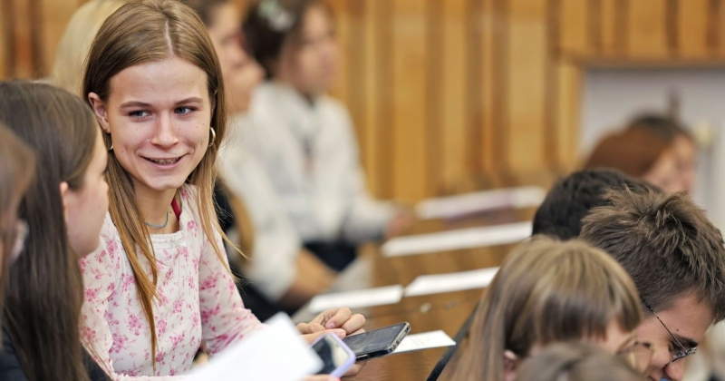 Studenci i studentki na sali wykładowej, przed nimi leżą kartki, po lewej stronie widać twarz dziewczyny, która rozmawia z koleżanką