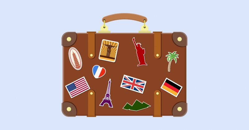 Grafika. Brązowa walizka z naklejkami. Naklejki to flagi lub znane budowle na całym świecie. 