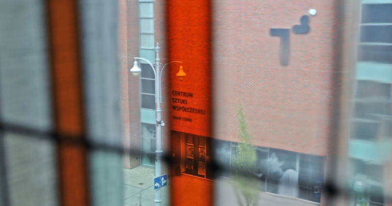 Widok na budynek CSW zza zamkniętego okna z budynku naprzeciwko. Napis na budynku "Centrum Sztuki Współczesnej Znaki Czasu" jest wyróżniony, bo widać go w pomarańczowej szybce witrażowego okna