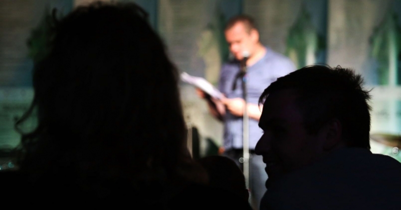 Na pierwszym planie widać głowy dwóch postaci, a w tle w oświetleniu na scenie mężczyzna stoi i czyta coś do mikrofonu z kartek, które trzyma