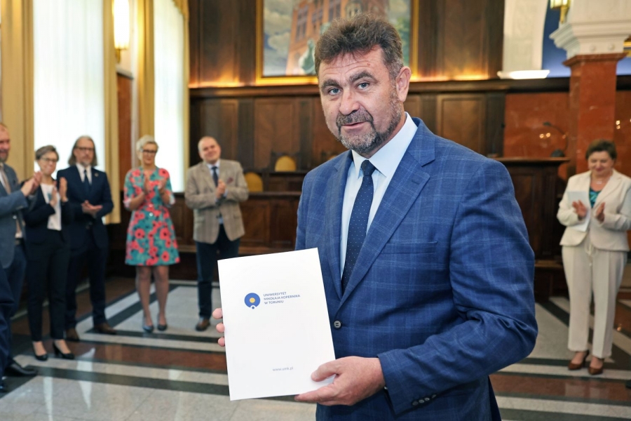Mężczyzna w średnim wieku, z krótko przystrzyżoną brodą, w ciemnoniebieskiej marynarce, pokazuje do obiektywu tekturową teczkę z aktem powołania; w tle klaszczące osoby; eleganckie wnętrze.