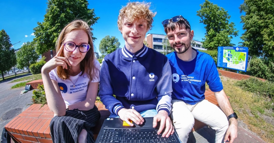Studentka i dwóch studentów siedzą obok siebie na dworze. Student siedzący na środku trzyma laptopa na kolanach. Studenci mają na sobie ubrania z logo UMK