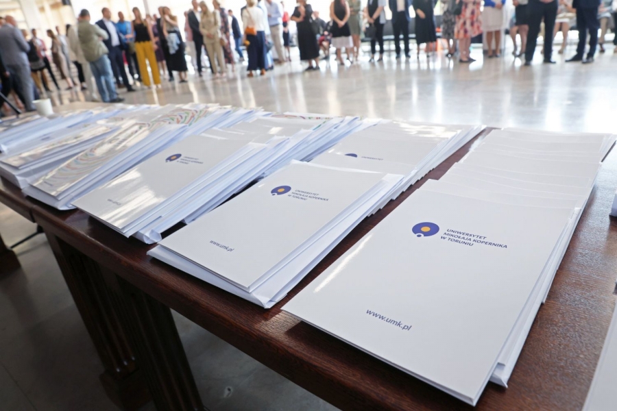 Na stole leżą stosy teczek z logo UMK. W głębi zdjęcia widać niewyraźne sylwetki osób