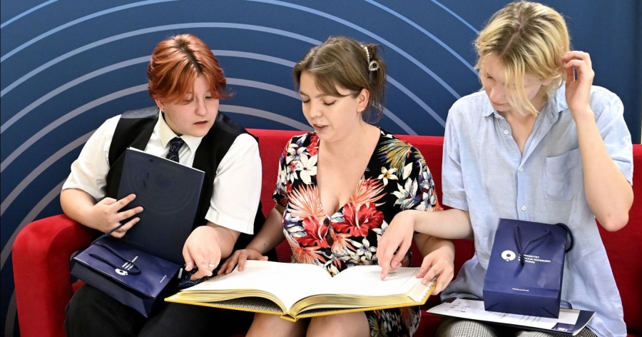 Trzy dziewczyny siedzą na czerwonej kanapie. Dziewczyna na środku trzyma na kolanach dużą otwartą książkę, dwie dziewczyny po bokach wskazują coś palcem w tekście