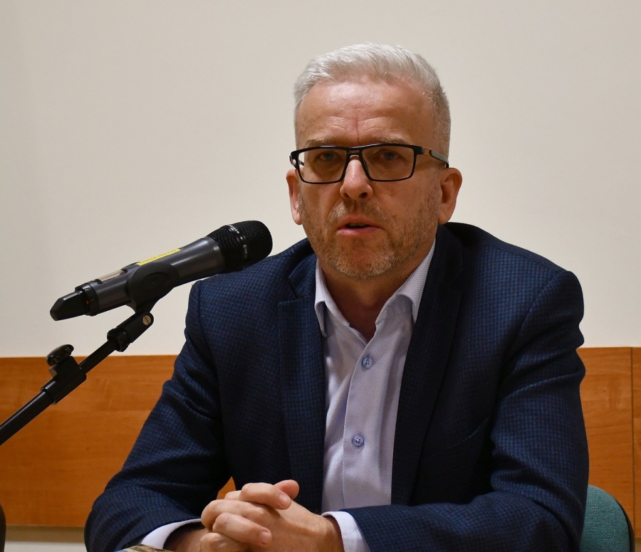 Siedzący mężczyzna w okularach i granatowej marynarce mówi do stojącego przed nim na stole mikrofonu.