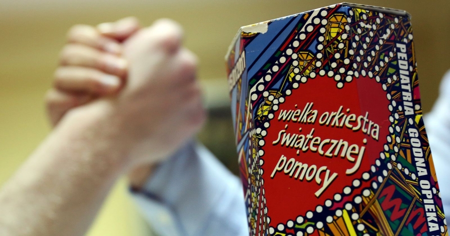 Na pierwszym planie widać puszkę WOŚP, a w tle dwie ręce ściskają się w przyjaznym geście