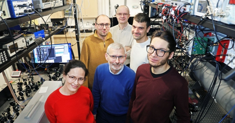 Grupa naukowców stoi w laboratorium. Widać sprzęt laboratoryjny. Ludzie są uśmiechnięci