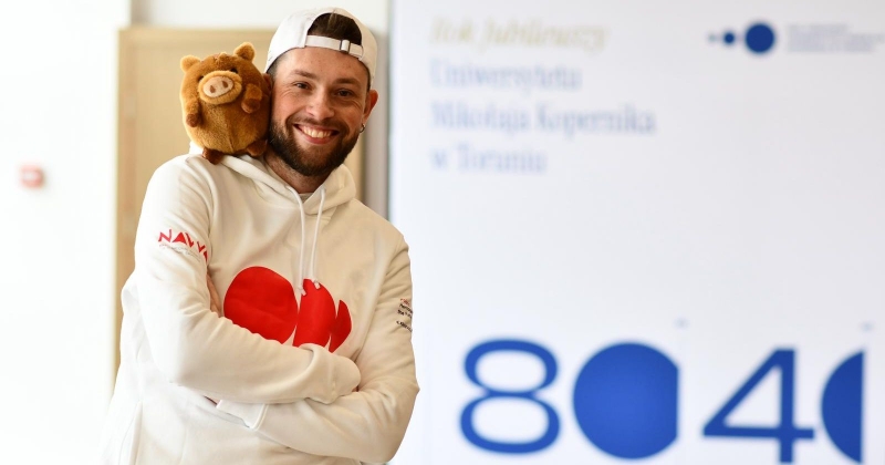 Mężczyzna w białej bluzie z czerwonym logo ptrojektu, na głowie ma czapkę z daszkiem, na ramieniu maskotkę