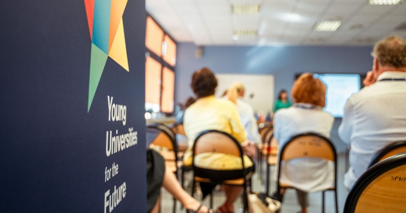 The photo shows people sitting backwards, listening to a lecture. On the left there is the navy blue roll-up with the YUFE logo