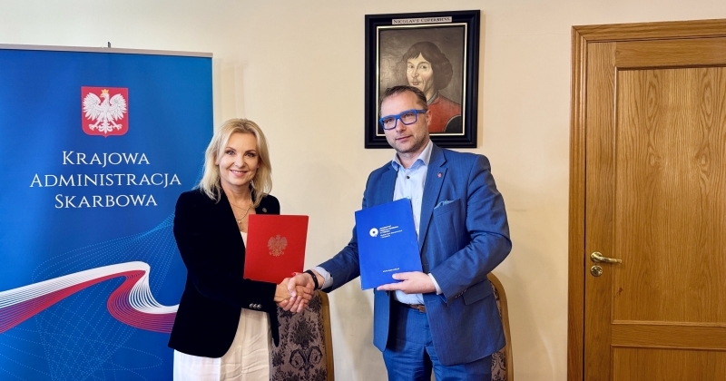 Dwoje ludzi, kobieta i mężczyzna, stoi w pomieszczeniu, podają sobie ręce i trzymają teczki ? kobieta czerwoną z godłem Polski, mężczyzna niebieską z logo instytucji. Za nimi widoczny jest niebieski baner z napisem ?Krajowa Administracja Skarbowa? oraz obraz przedstawiający Mikołaja Kopernika. Po prawej stronie widać drewniane drzwi.
