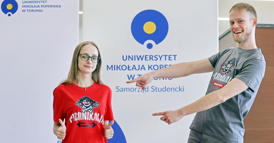 Marta Kuta i Hubert Długołęcki w koszulkach przygotowanych na tegoroczne Piernikalia. Hubert wskazuje palcami na Martę. W tle stoi ścianka z logo Samorządu Studenckiego