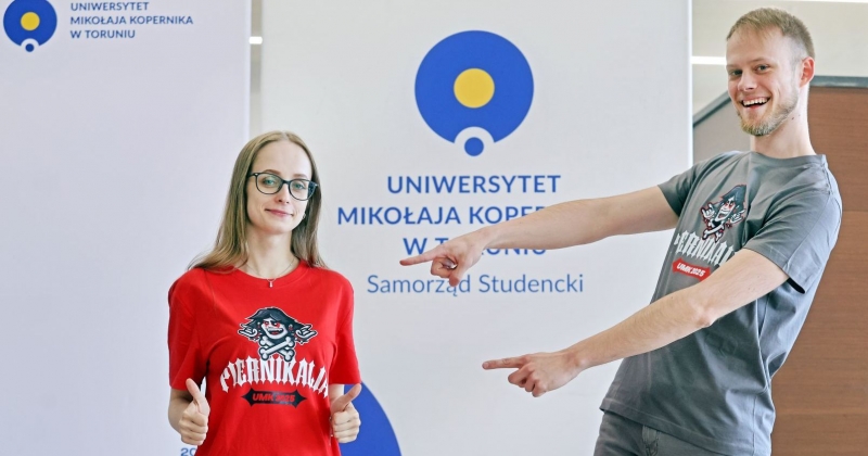 Marta Kuta i Hubert Długołęcki w koszulkach przygotowanych na tegoroczne Piernikalia. Hubert wskazuje palcami na Martę. W tle stoi ścianka z logo Samorządu Studenckiego