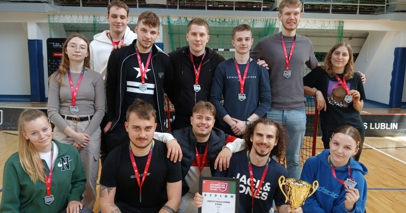  Grupa trzynastu młodych osób - zawodniczek i zawodników z drużyny unihokeja Uniwersytetu Mikołaja Kopernika w Toruniu - pozuje na hali sportowej. Wszyscy mają na szyjach srebrne medale, stoją i klęczą w dwóch rzędach. Jedna osoba trzyma puchar i dyplom. 