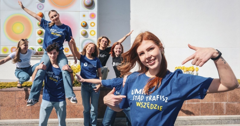 Grupka uśmiechniętych studentów i studentek w koszulkach z hasłem "UMK stąd trafisz wszędzie", w tle mozaika Stefana Knappa zdobiąca fasadę Auli UMK