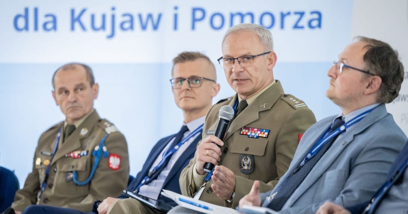 Zdjęcie czterech mężczyzn podczas panelu dyskusyjnego. Dwóch ma na sobie mundury wojskowe, a pozostali garnitury. Jeden z umundurowanych mężczyzn trzyma mikrofon i przemawia. W tle fragment baneru z napisem "dla Kujaw i Pomorza"