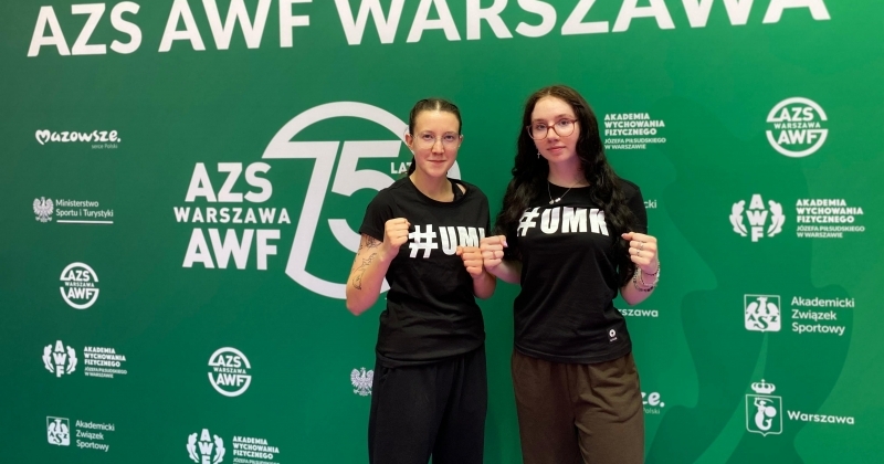 ?Dwie osoby w czarnych koszulkach z napisem #UMK stoją na tle zielonej ścianki z logotypami i napisami, w tym 'AZS AWF WARSZAWA', '75 LAT', 'Ministerstwo Sportu i Turystyki', 'Akademia Wychowania Fizycznego' oraz 'Akademicki Związek Sportowy'.