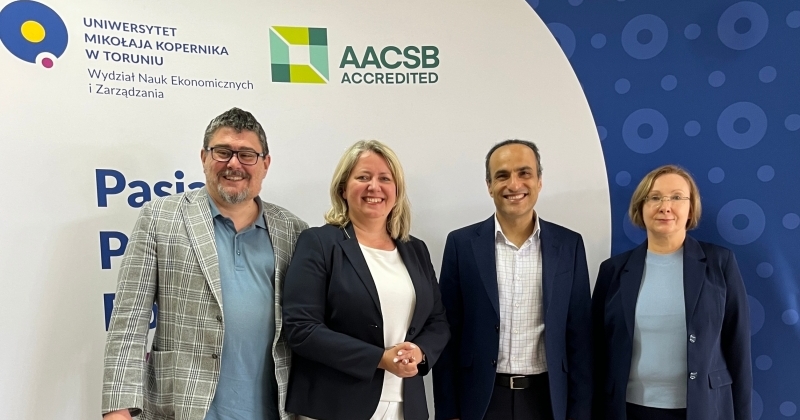 3 nauczycieli akademickich WNEiZ UMK i profesor z Australii stoją na tle logo wydziału i logo AACSB.