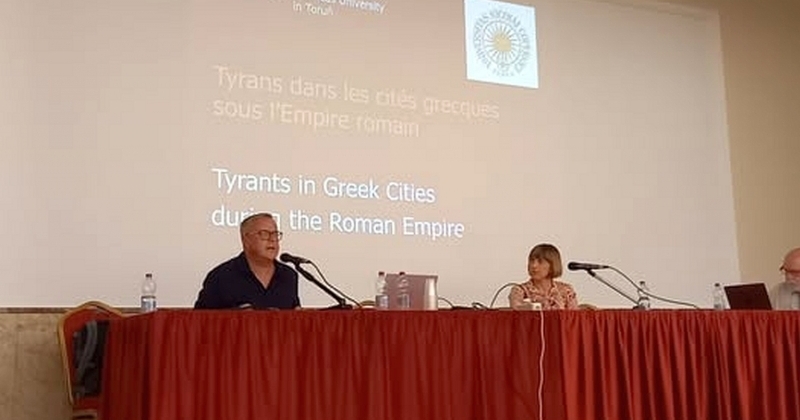 Dwójka osób - mężczyzna i kobieta - siedzą na podwyższeniu za stołem przykrytym czerwonym suknem; w tle ekran z tytułowym slajdem prezentacji.