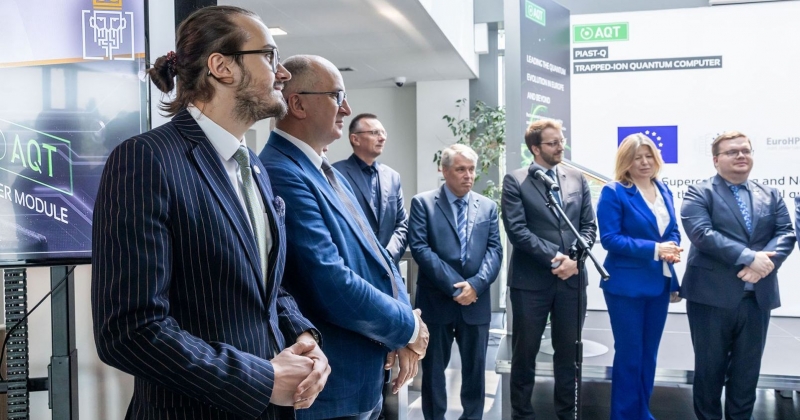 Siedem elegancko ubranych osób stoi jedna obk drugiej podczas otwarcia fabryki. W tle prezentacja, widać flagę Unii Europejskiej